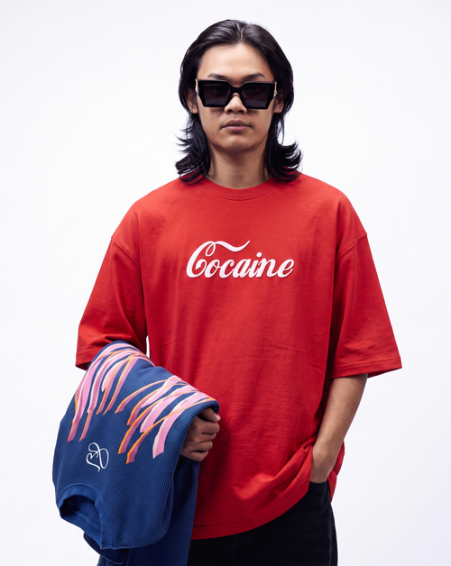 A model wearing a red oversized t-shirt with 'Cocaine' printed in white script across the chest, holding a blue t-shirt with pink and orange graphic stripes. The model is wearing black sunglasses and black pants against a plain white studio background.